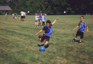 Kids playing kickball on a grassy field wearing blue pinnies at YMCA Camp Lowe in Central Massachusetts