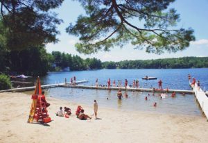 Children swimming and playing at a lakeside beach with a lifeguard at YMCA Camp Lowe in Central Massachusetts