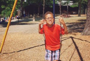 Child wearing a red shirt sitting on a swing in a playground area at YMCA Camp Lowe in Central Massachusetts