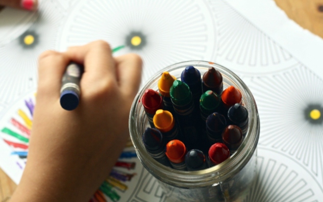 A child is using a blue crayon to color next to a glass jar filled with colorful crayons